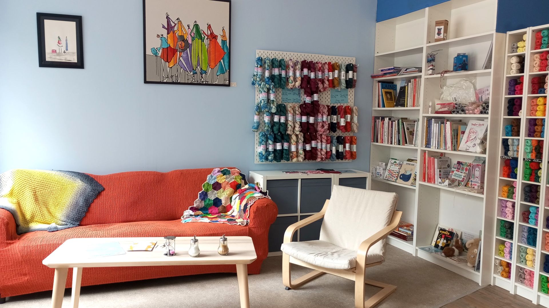 Photo d'un salon de thé avec canapé orange, des laines en exposition sur le mur et une bibliothèque de livres sur le crochet et le tricot